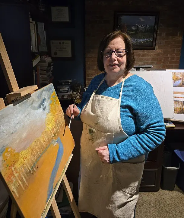 A woman in a blue shirt and apron stands beside an easel, crafting original canvas art with a brush as she paints a landscape of trees and a river.